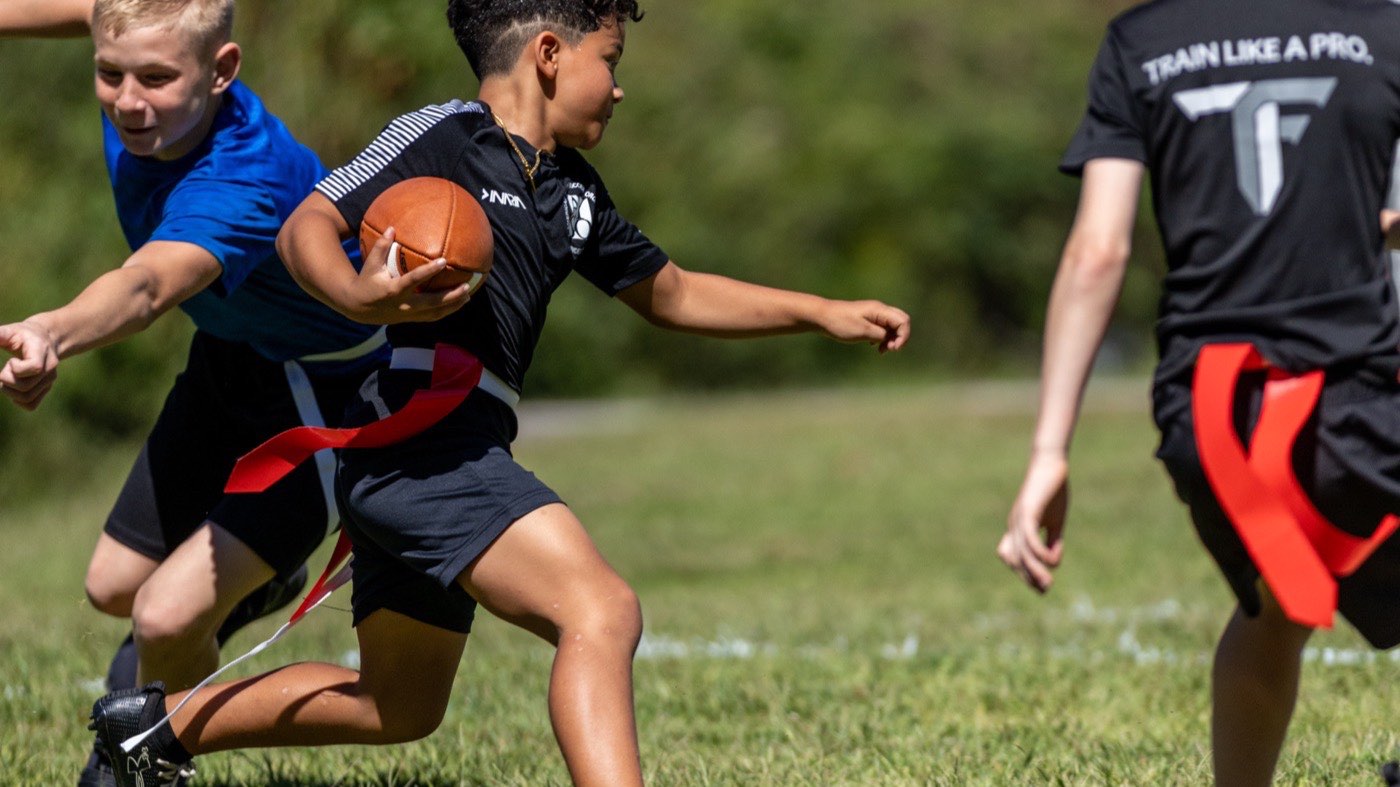 A young player carries the ball past a defender reaching for his flag.