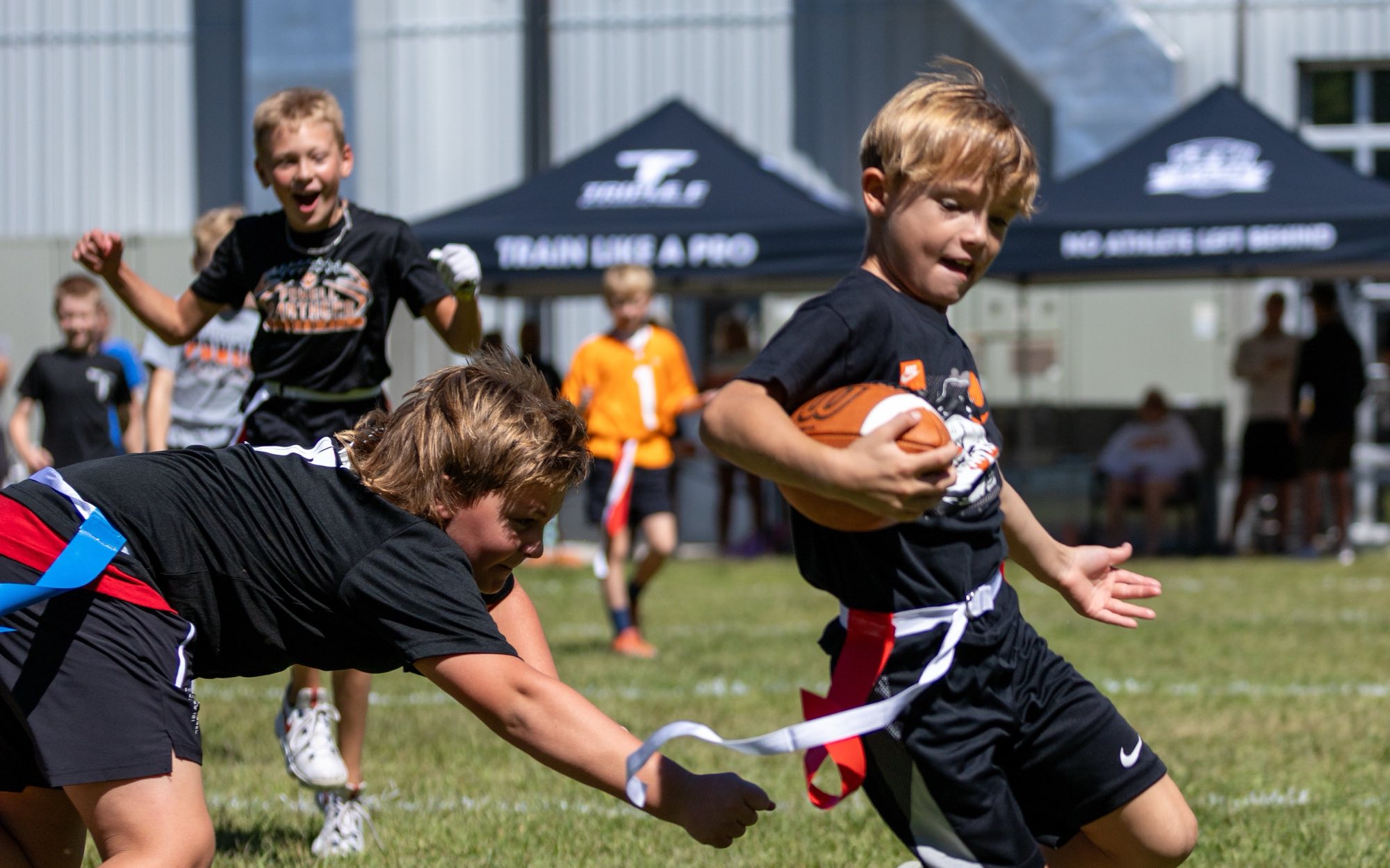 A young flag football player carries the ball past a diving defender reaching for his flag, with team tents behind the field.