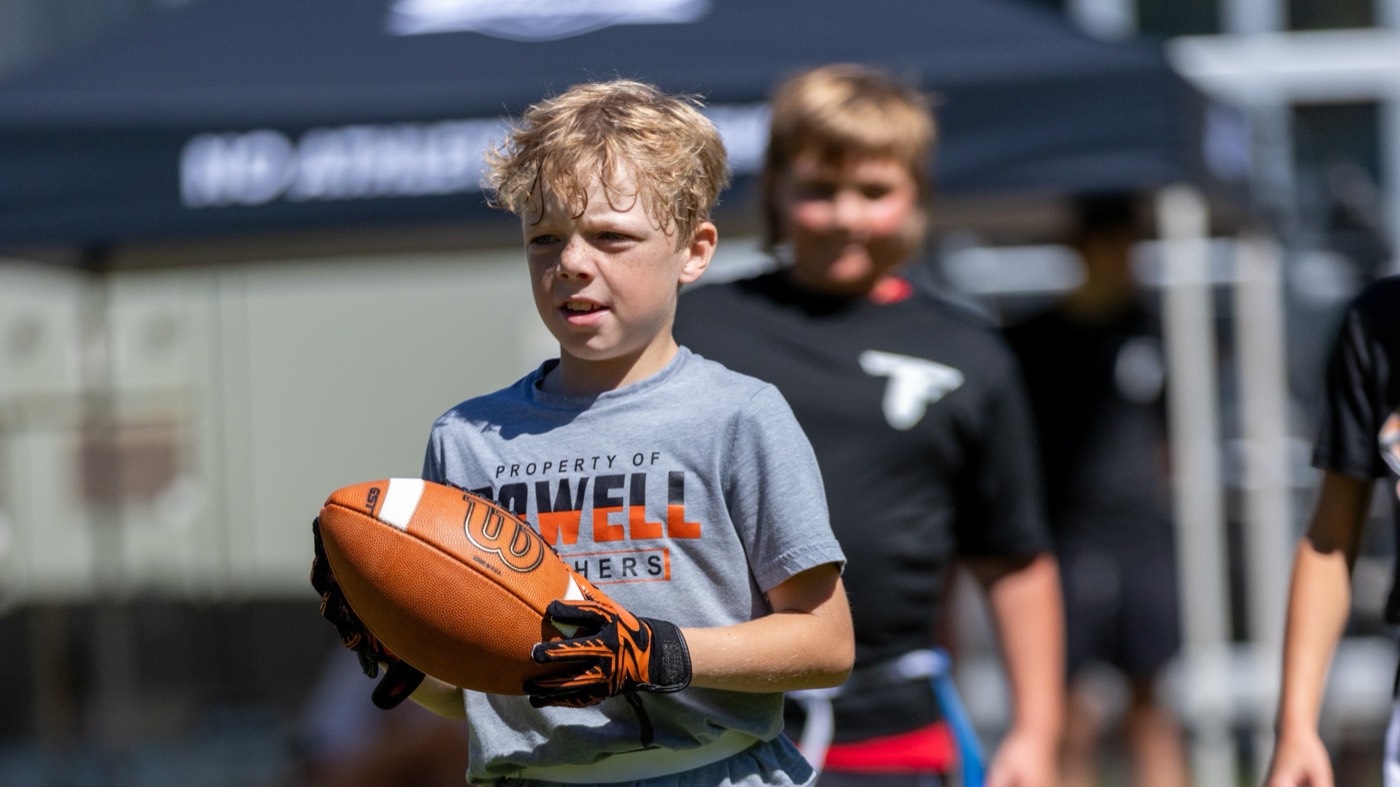 A young player smiles as he walks off the field with the ball, teammates and a team tent behind him.